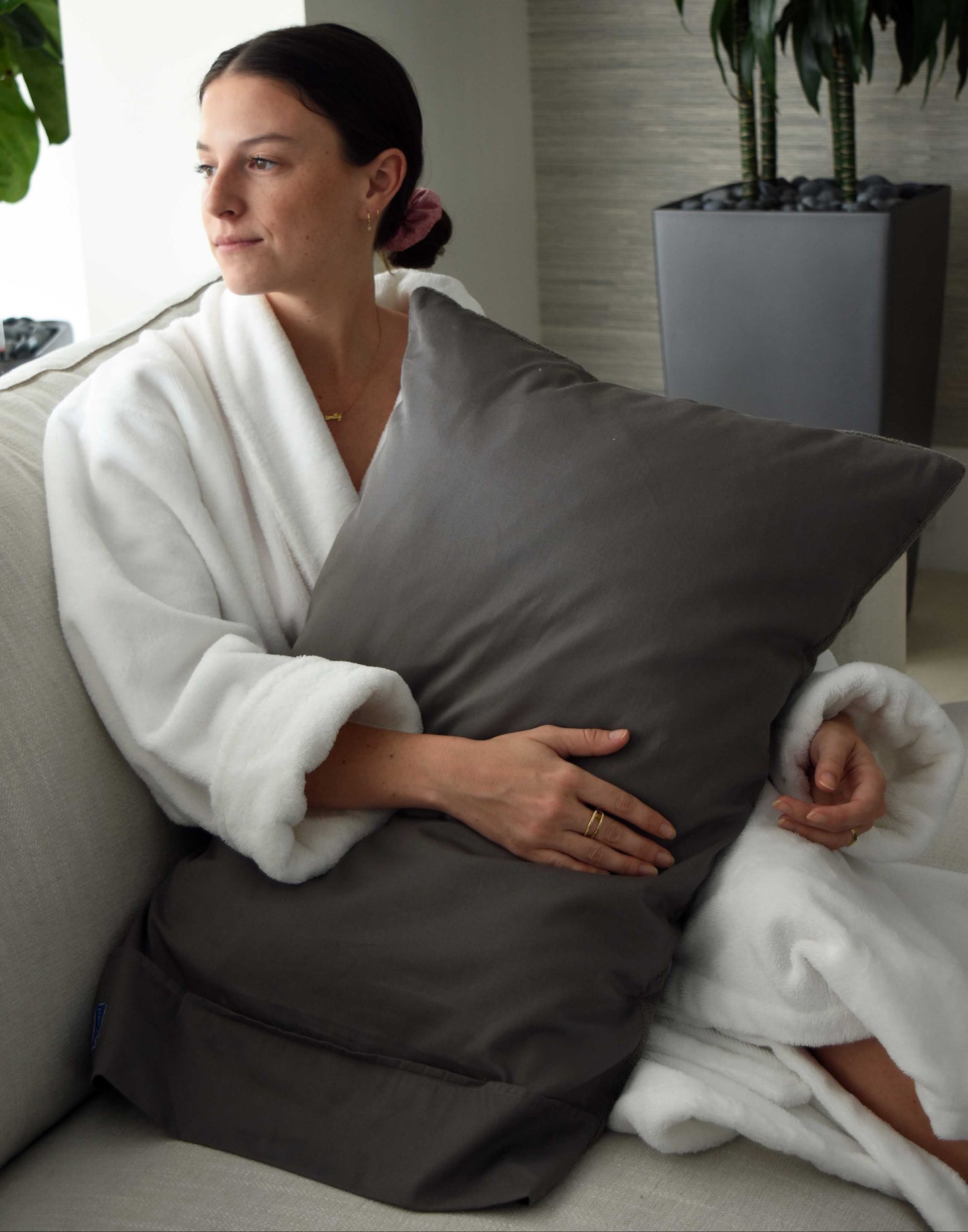 Woman in a white robe holding a gray pillow in a room with plants.