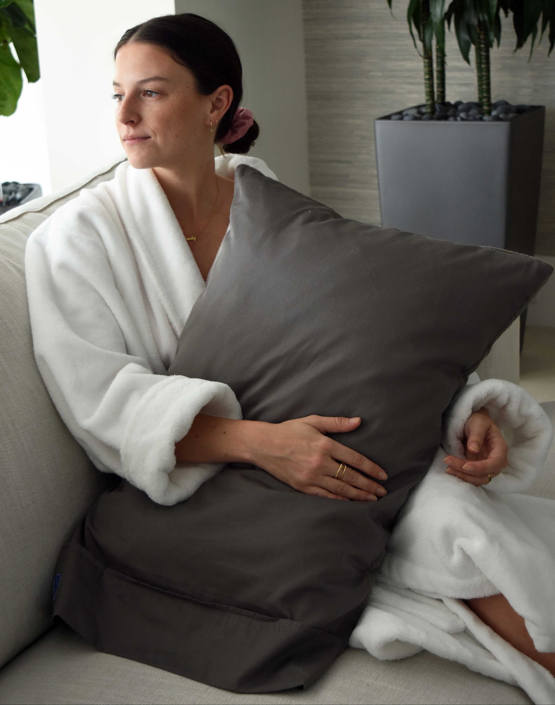 Woman in a white robe holding a gray pillow in a room with plants.
