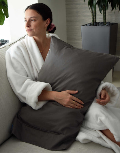 Woman in a white robe holding a gray pillow in a room with plants.
